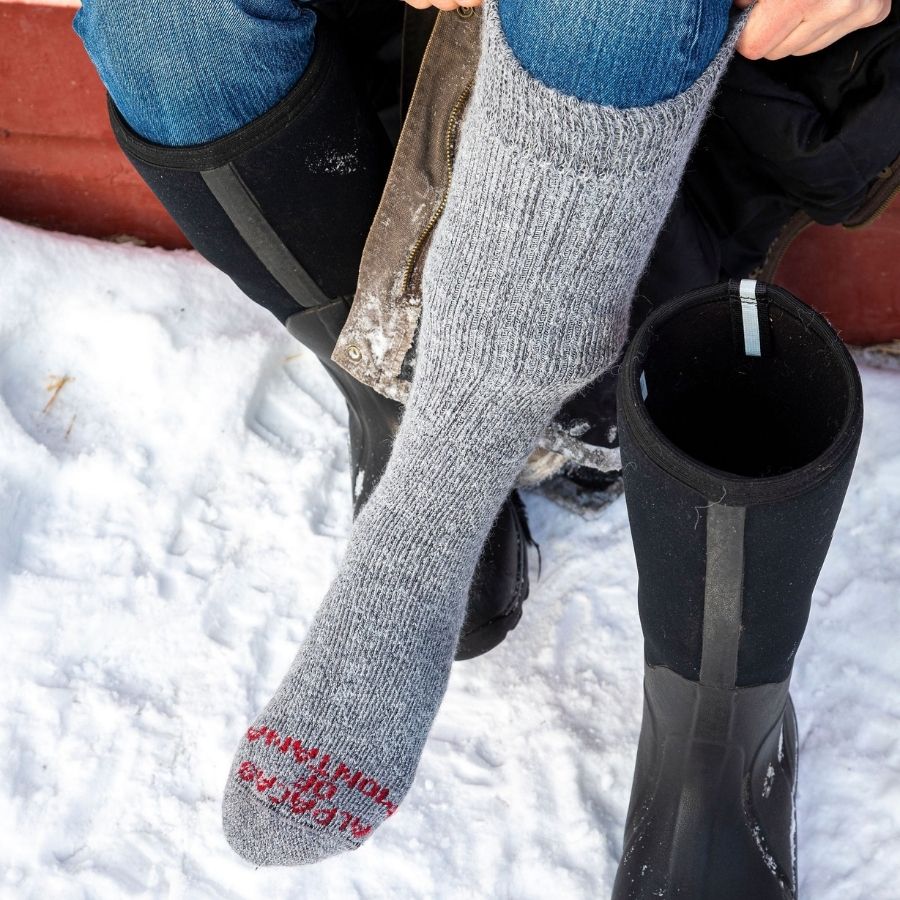 A person sitting on wooden steps with a tall boot on one foot and pulling up a sock on the other. The sock is the silver gray Alpacas of Montana cozy comfortable soft warm thermal winter freezing temperatures moisture wicking maximum warmth arctic boot socks for hiking, snowshoeing, hunting, skiing, ice fishing, outdoors.