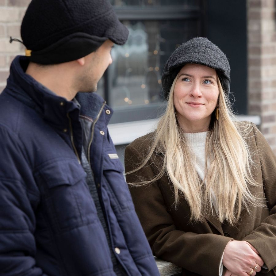 A blonde woman smiling wearing gray extremely warm cozy soft windproof thermal alpaca fleece wool city commuter winter hat looking at a man wearing extremely warm black cozy comfortable soft moisture wicking winter alpaca wool city commuter hat