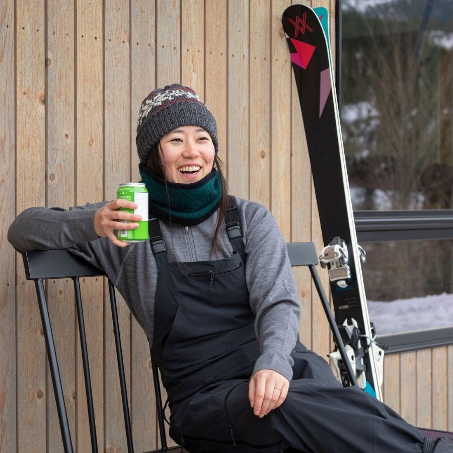 A smiling woman with black hair sitting on a metal bench next to a pair of skis and holding a drink can. She is wearing black overall snowpants, a gray Alpacas of Montana women&#39;s base layer top, a knitted hat, and an Alpacas of Montana lightweight cozy comfortable moisture wicking antimicrobial warm breathable all seasons thin thermal outerwear skiing hiking climbing outdoors hunting fishing rocky mountain neck gaiter made from dark forest green and black alpaca wool.