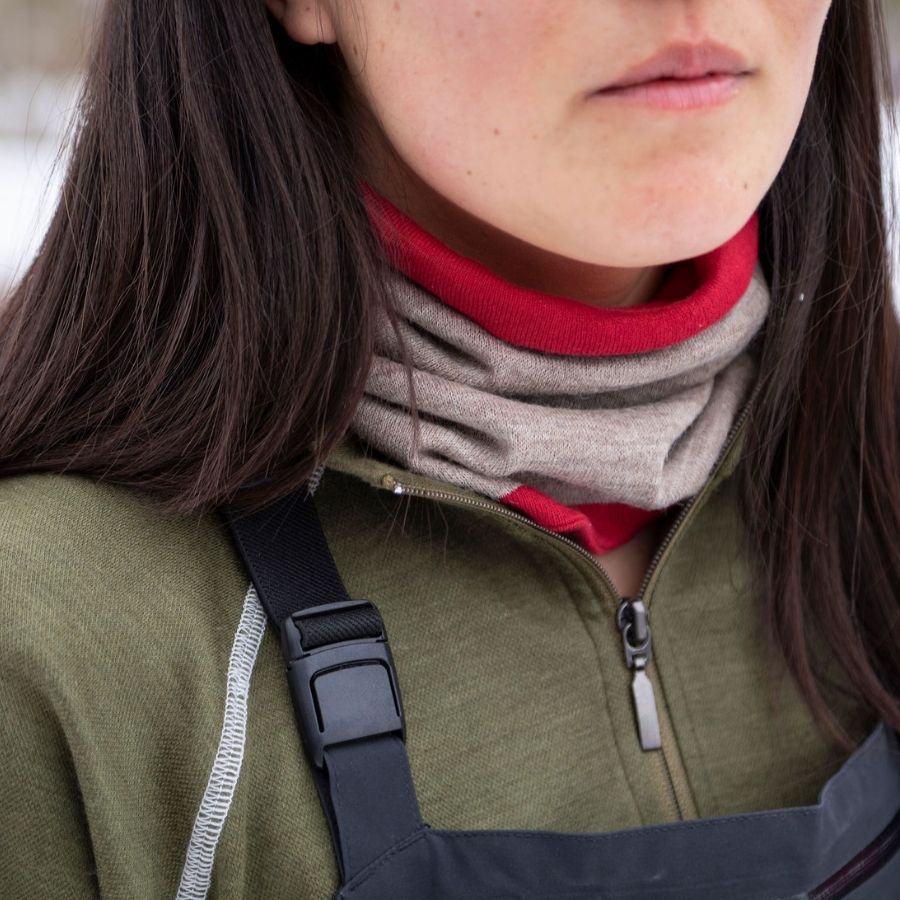 A close up photo of a black haired woman wearing black overall snowpants, olive green Alpacas of Montana women&#39;s base layer top, and a lightweight cozy comfortable moisture wicking warm breathable all seasons thin thermal outerwear skiing hiking climbing outdoors hunting fishing high desert neck gaiter made from scarlet red and light camel brown alpaca.