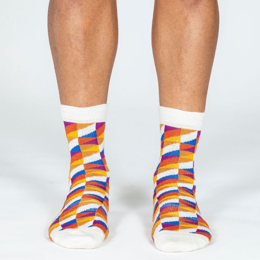 A person&#39;s lower legs standing against a white background wearing a pair of Alpacas of Montana white, yellow, gold, orange, purple, and blue triangle pattern casual lounge fashion comfortable soft cozy everyday moisture wicking alpaca wool Swag socks.