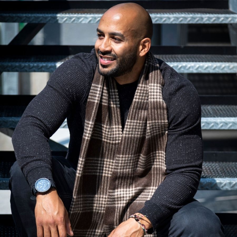 A bald man with a black beard looking to the side and sitting on a set of metal stairs. He is wearing black jeans, a speckled black long sleeve shirt, and an Alpacas of Montana soft stylish men&#39;s fashion comfortable cozy warm alpaca wool chocolate brown and latte brown plaid pattern scarf with tassels.
