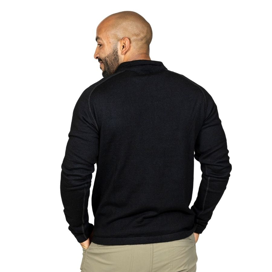 A smiling bald man with a black beard standing facing away from the camera with his hands in his pockets in front of a white background. He is wearing khaki pants and a black Alpacas of Montana warm thermal soft cozy comfortable activewear outerwear athletic moisture wicking antimicrobial men&#39;s fashion stylish luxury mid-layer quarter-zip alpaca wool long sleeve pullover top for outdoors camping climbing hiking skiing hunting fishing running winter.