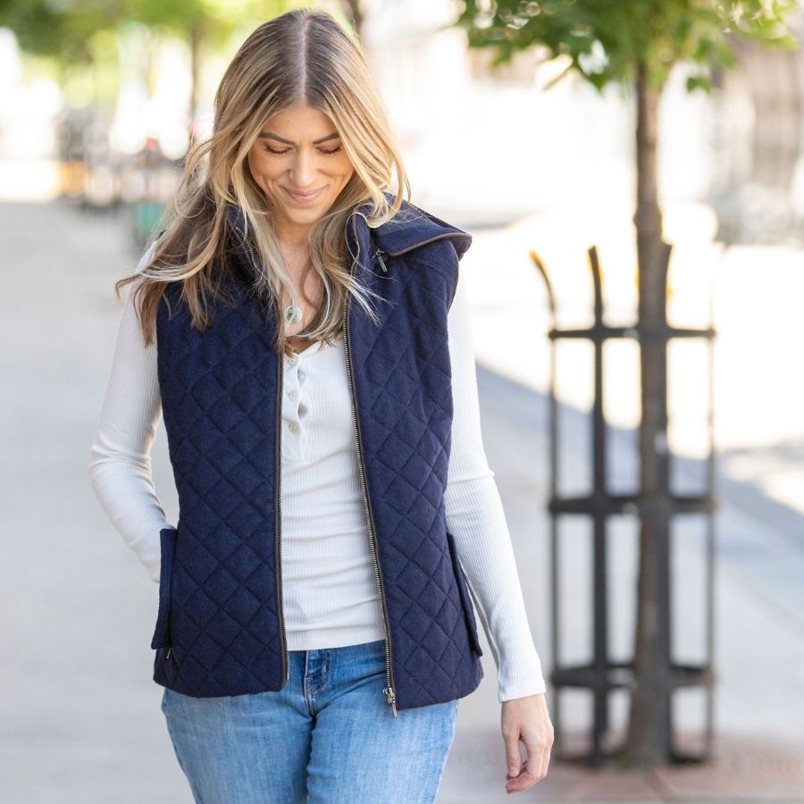 A blonde woman walking on a sidewalk and smiling at the ground. She is wearing blue jeans, a white longsleeve shirt, and a dark navy ocean blue Alpacas of Montana soft comfortable cozy casual fancy luxury fashionable stylish cozy thermal moisture wicking stretchy quilted hooded women&#39;s fitted alpaca wool vest.
