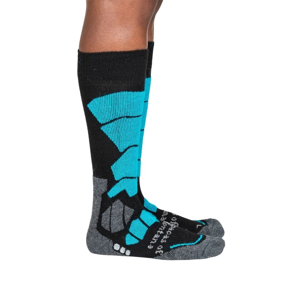 A person&#39;s lower legs standing sideways with a white background wearing teal bue, gray, and black Alpacas of Montana cozy comfortable soft warm thermal winter freezing temperatures antimicrobial moisture wicking alpaca wool ski and snowboard socks for snowshoeing, skiing, snowboarding, ice fishing, outdoors.