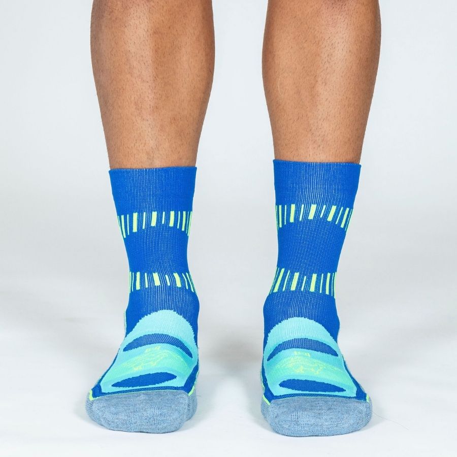 A photo of a person&#39;s lower legs standing against a white background wearing cobalt royal blue, sky blue, and lime yellow Alpacas of Montana soft cozy comfortable activewear outerwear athletic workout moisture wicking antimicrobial cushioned light compression engineered high-tech mid-crew hiking sock for walking, skiing, climbing, hunting, camping, fishing, exercise, biking