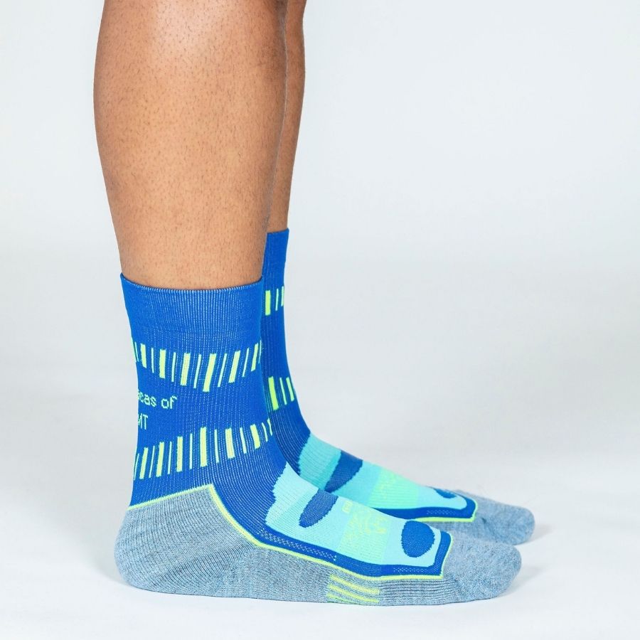 A photo of a person&#39;s lower legs standing sideways to the camera against a white background wearing cobalt royal blue, sky blue, and lime yellow green Alpacas of Montana soft cozy comfortable activewear outerwear athletic workout moisture wicking antimicrobial cushioned light compression engineered high-tech mid-crew hiking sock for walking, skiing, climbing, hunting, camping, fishing, exercise, biking
