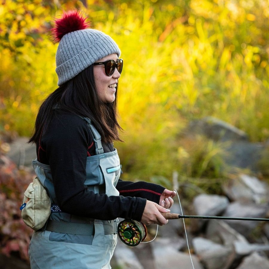 woman smiling while fly fishing and wearing a soft warm winter cozy moisture wicking comfortable fashionable light gray alpaca wool beartooth beanie with ruby red pom pom