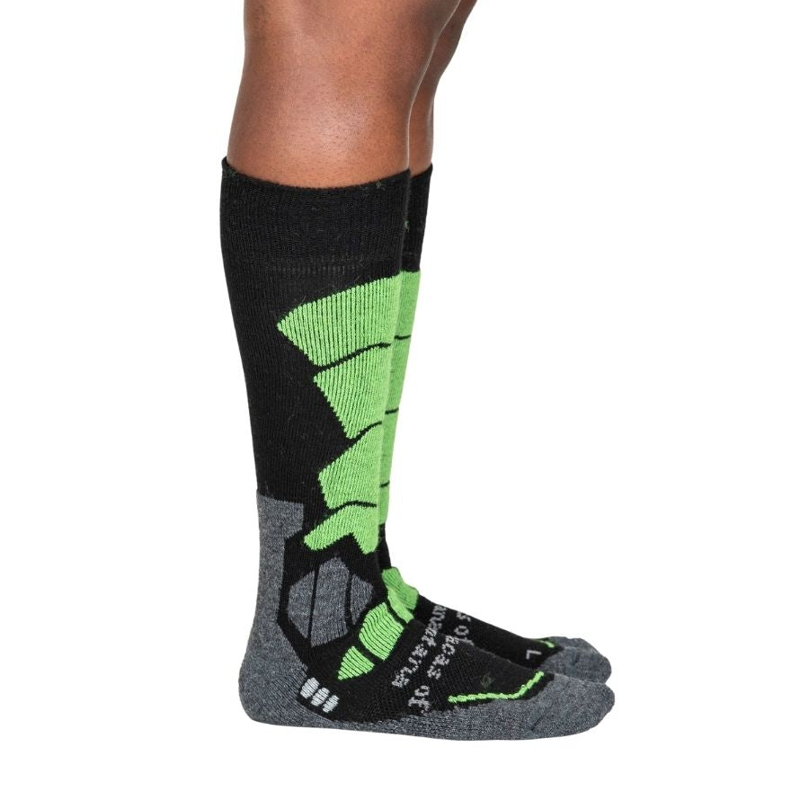 A person&#39;s lower legs standing sideways with a white background wearing lime green, gray, and black Alpacas of Montana cozy comfortable soft warm thermal winter freezing temperatures antimicrobial moisture wicking alpaca wool ski and snowboard socks for snowshoeing, skiing, snowboarding, ice fishing, outdoors.