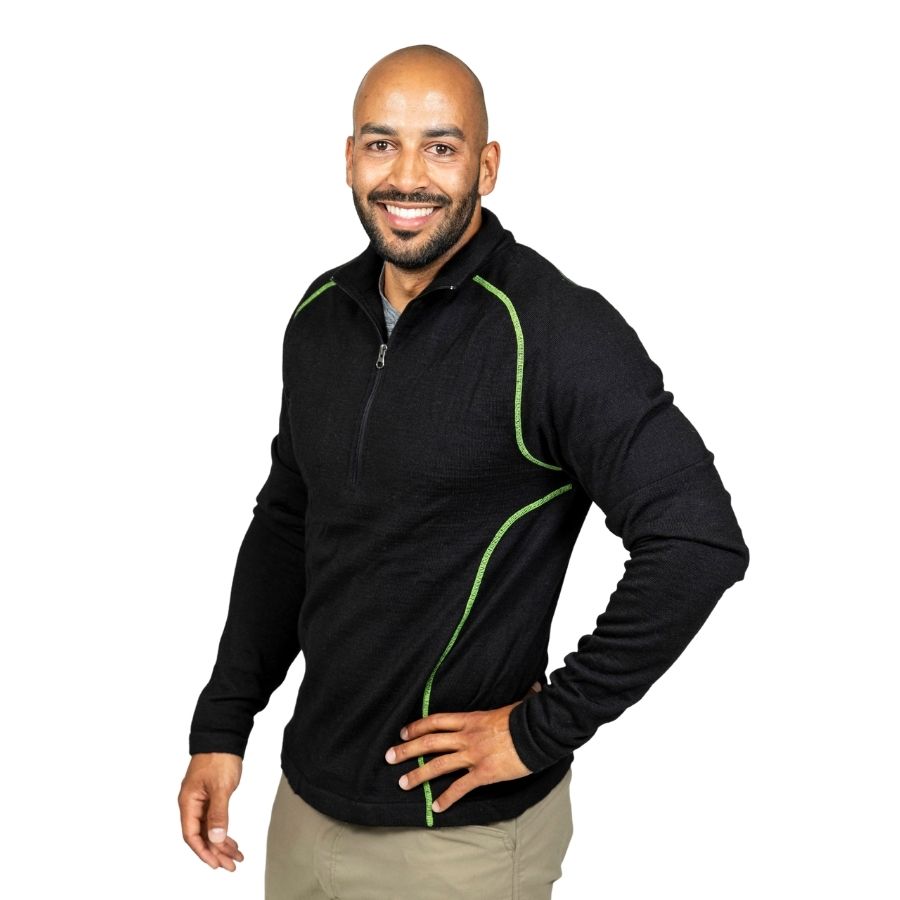 A smiling bald man with a black beard standing with one hand on his hip in front of a white background. He is wearing khaki pants and a black with lime green stitching Alpacas of Montana warm thermal soft cozy comfortable activewear outerwear athletic moisture wicking antimicrobial men&#39;s fashion stylish luxury mid-layer quarter-zip alpaca wool long sleeve pullover top for outdoors camping climbing hiking skiing hunting fishing running winter.