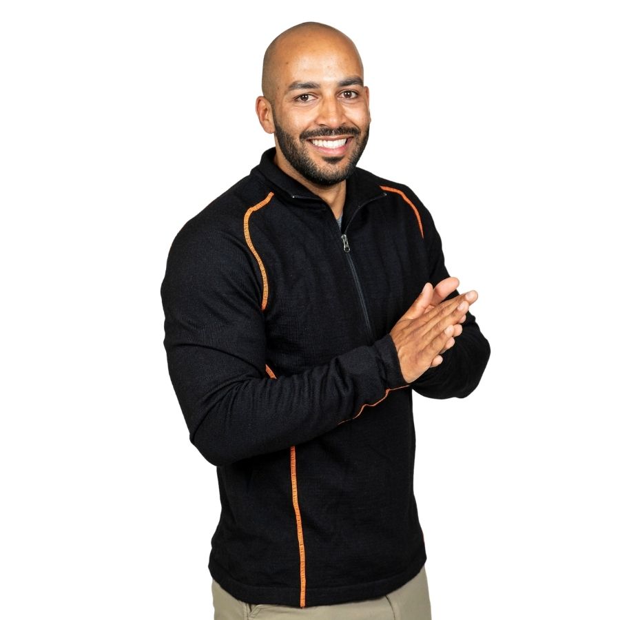 A smiling bald man with a black beard standing in front of a white background. He is wearing khaki pants and a black with orange stitching Alpacas of Montana warm thermal soft cozy comfortable activewear outerwear athletic moisture wicking antimicrobial men&#39;s fashion stylish luxury mid-layer quarter-zip alpaca wool long sleeve pullover top for outdoors camping climbing hiking skiing hunting fishing running winter.