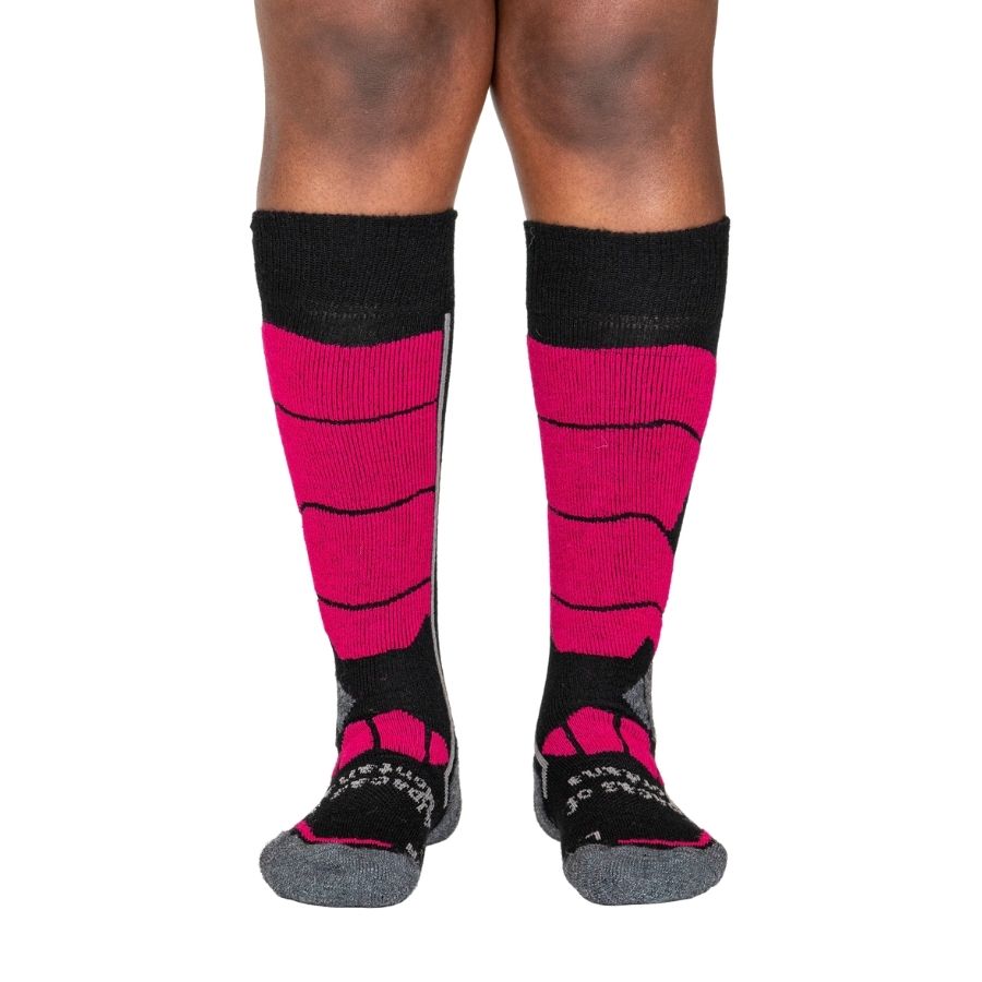 A person&#39;s lower legs with a white background wearing fuchsia pink, gray, and black Alpacas of Montana cozy comfortable soft warm thermal winter freezing temperatures antimicrobial moisture wicking alpaca wool ski and snowboard socks for snowshoeing, skiing, snowboarding, ice fishing, outdoors.