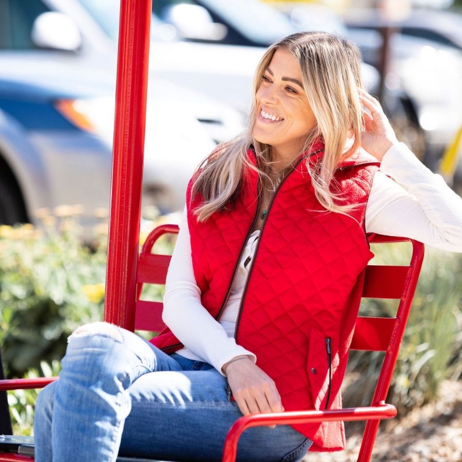A smiling blonde woman sitting in a red ski chairlift seat and looking off to her right. She is wearing blue jeans, a white long sleeve shirt, and a bright cherry scarlet red Alpacas of Montana soft comfortable cozy casual fancy luxury fashionable stylish cozy thermal moisture wicking stretchy quilted hooded women&#39;s fitted alpaca wool vest.