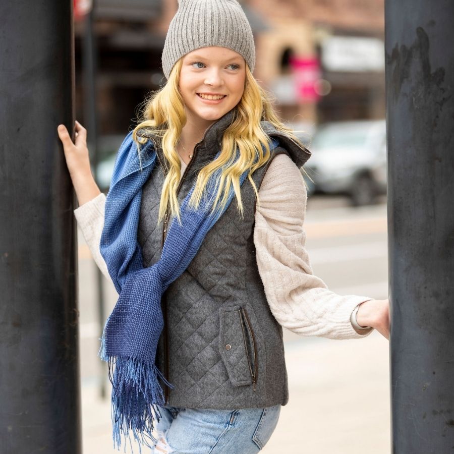 A blonde woman smiling at something to her left between two street lamp poles. She is wearing a light gray Alpacas of Montana beartooth beanie, a drifting blues Alpacas of Montana shawl, blue jeans, a fluffy white long sleeve shirt, and a neutral mid gray Alpacas of Montana soft comfortable cozy casual fancy luxury fashionable stylish cozy thermal moisture wicking stretchy quilted hooded women&#39;s fitted alpaca wool vest.