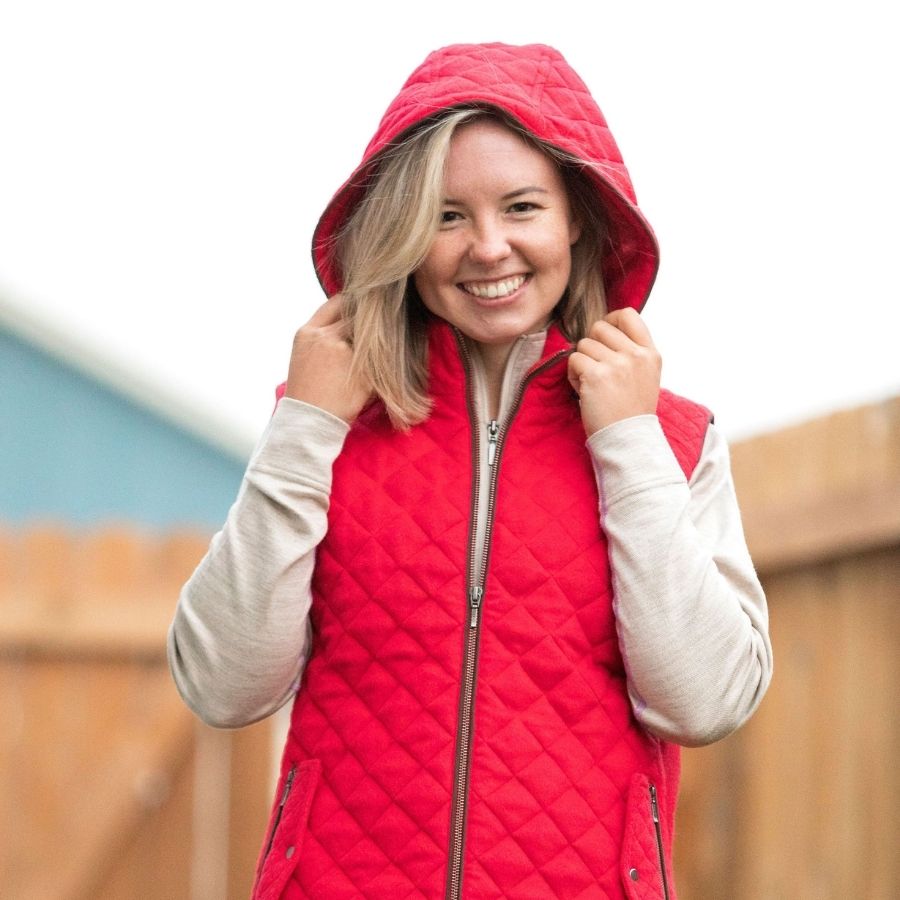 A smiling blonde woman standing in front of a wooden fence with her hands on her hood. She is wearing an oatmeal color Alpacas of Montana women&#39;s base layer top and a bright cherry scarlet red Alpacas of Montana soft comfortable cozy casual fancy luxury fashionable stylish cozy thermal moisture wicking stretchy quilted hooded women&#39;s fitted alpaca wool vest.