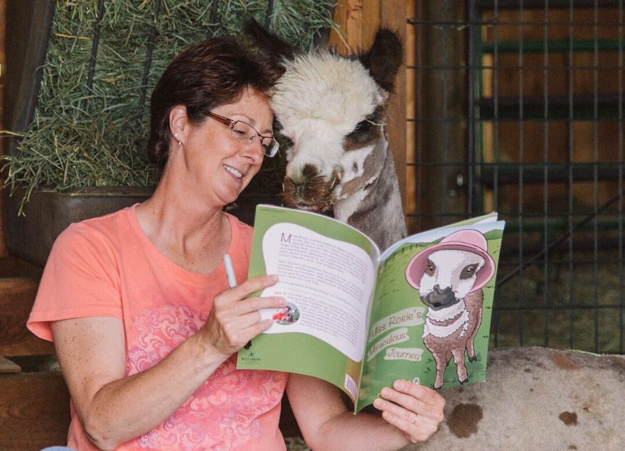 woman sitting in front of hay bail reading to an alpaca