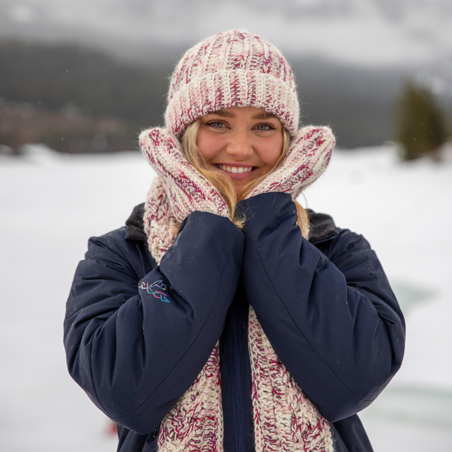 Smiling blonde haired woman standing in a winter scene wearing the dark pink, light pink, carnation, and white matching alpaca and bamboo soft scarf, hat, and mitten set from Alpacas of Montana. 
