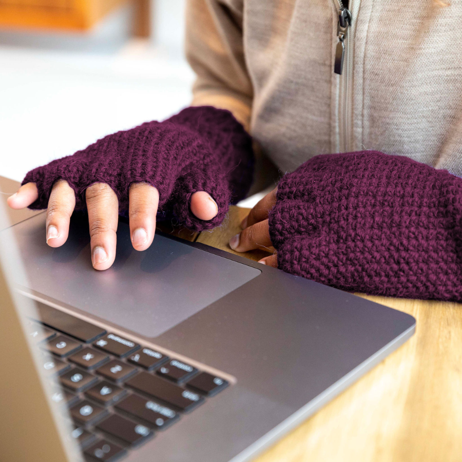 A close up photo of a pair of hands touching a laptop track pad wearing a pair of soft cozy comfortable fashionable moisture wicking knitted crochet fingerless gloves handmade in Montana from deep purple alpaca wool yarn.