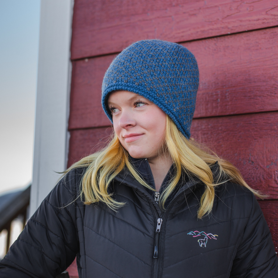 A blonde woman standing in front of a red barn wall and looking off to her right. She is wearing a black Alpacas of Montana women's tempest lite jacket and a blue and gray Alpacas of Montana extremely warm cozy soft windproof comfortable antimicrobial moisture wicking thermal alpaca fleece wool winter fleece lined beanie hat for hiking, skiing, snowshoeing, sledding, outdoors.