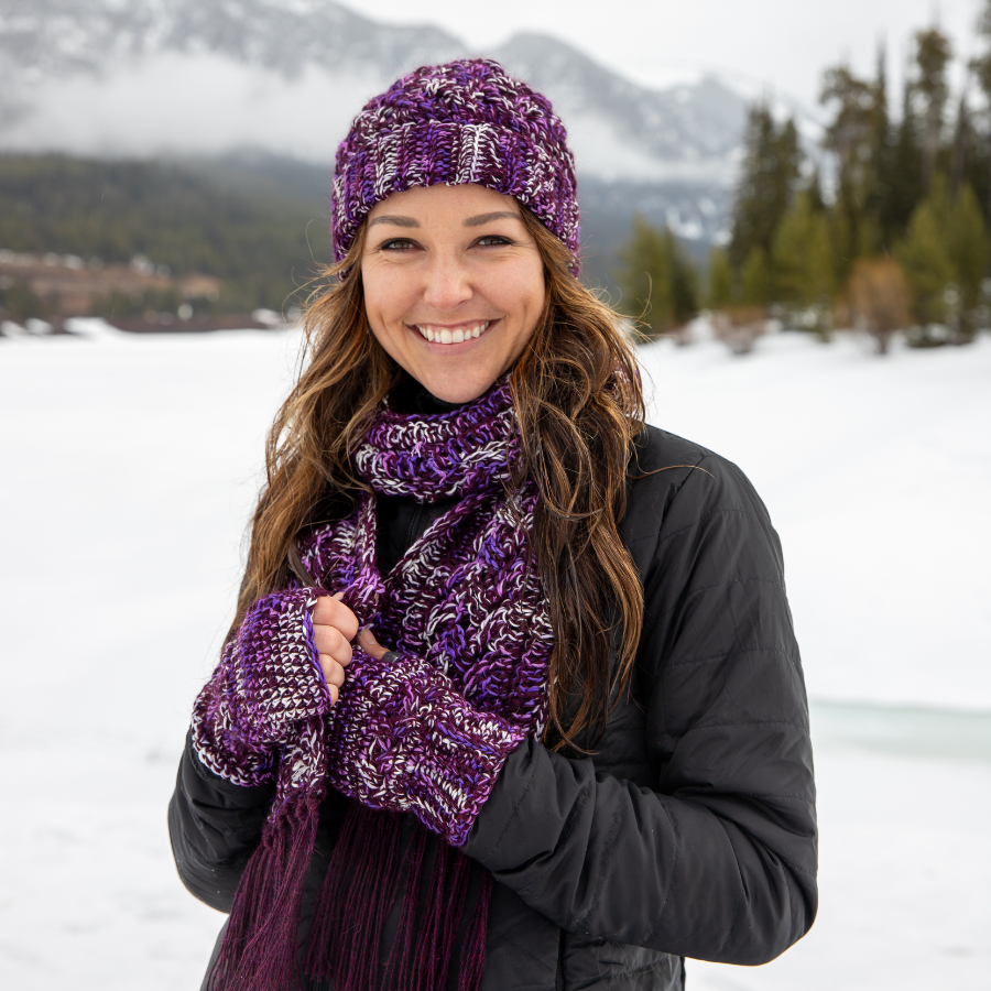 Smiling woman with brown hair standing in winter scene wearing Alpacas of Montana purple knitted crochet handmade matching set of hat, scarf, and fingerless wristie mittens. Handmade items are a mixture of deep purple, bright purple violet, and white with long tassels at the end of the scarf.