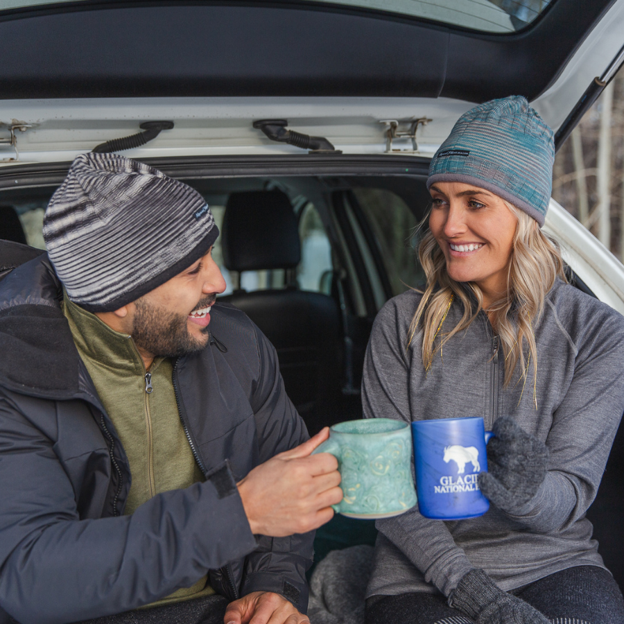 Man with black beard and woman with blonde hair sitting in the back of a car holding mugs. The man is wearing Alpacas of Montana charcoal color Granite Peak Parka, olive green Men's Base Layer Top, and soft cozy warm outerwear black, white, and gray Backcountry Beanie. The woman is wearing a gray Women's Base Layer Top and a soft cozy warm moisture wicking hiking skiing teal and gray Backcountry Beanie.