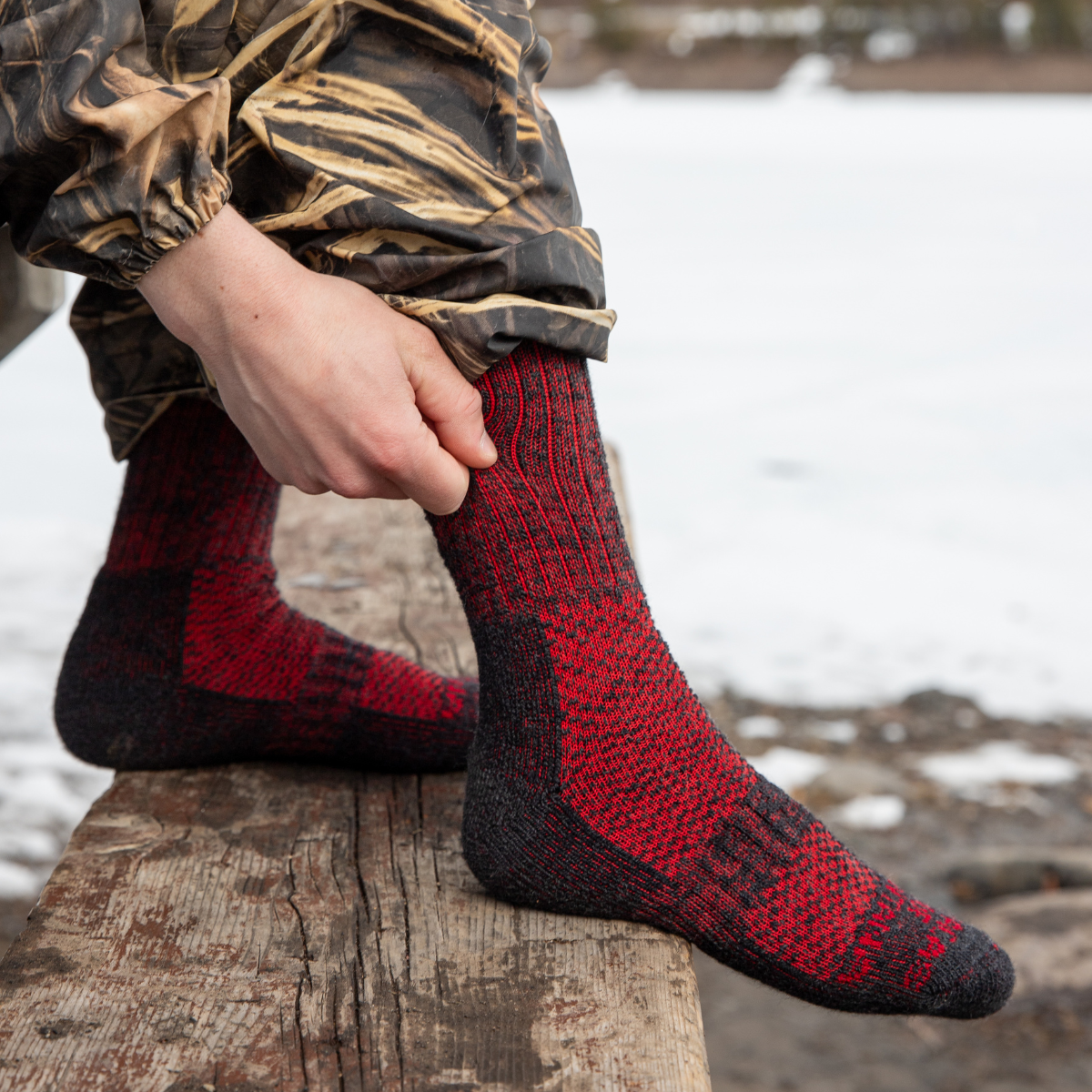 A close up of a hunter wearing camouflage sitting on a bench and pulling up a pair of Alpacas of Montana cozy soft warm comfortable thermal moisture wicking everyday winter fishing hiking snowshoeing hunting outdoors scarlet and black wine red extra cushion boot socks.