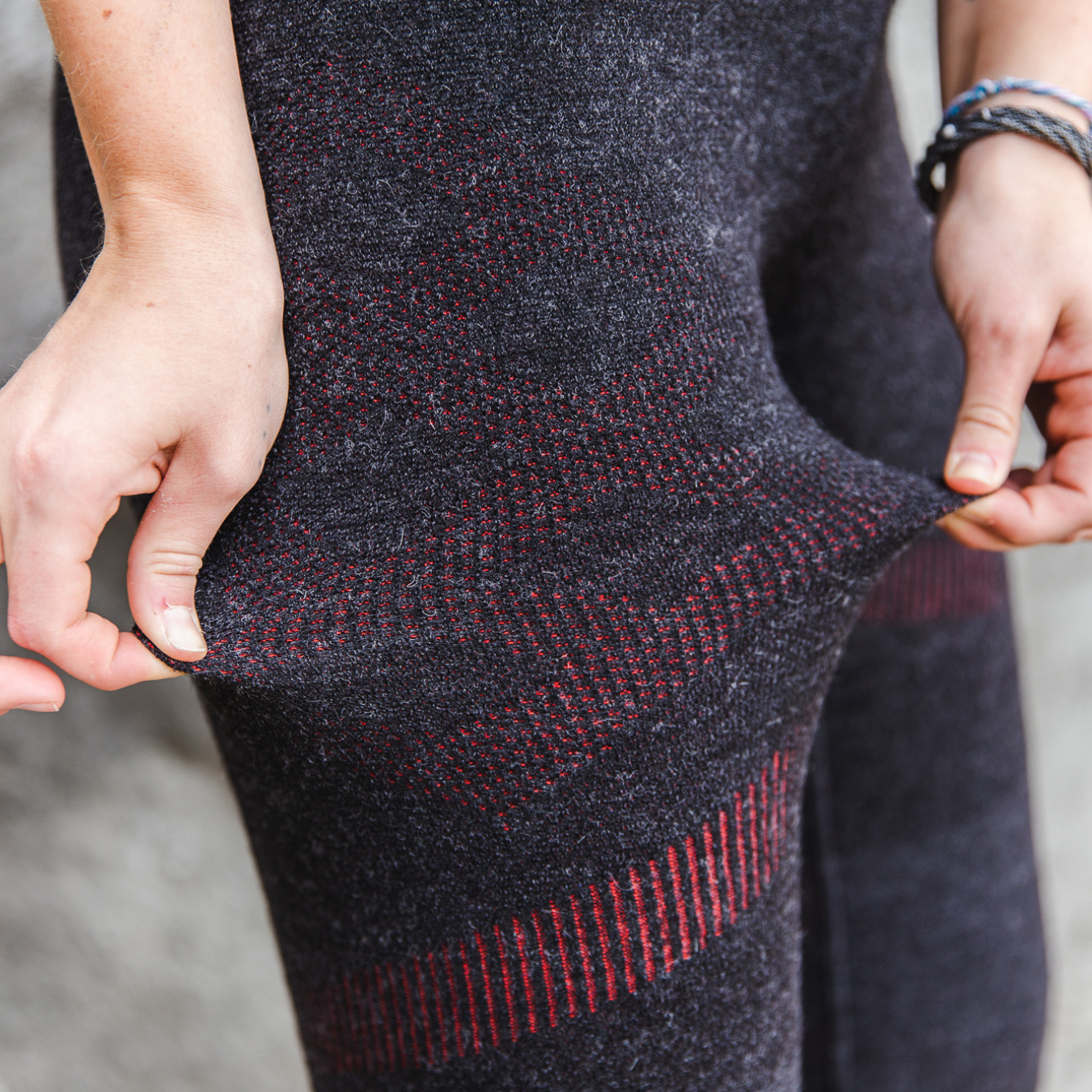  A close up photo of a pair of hands demonstrating the stretch capabilities of the Alpacas of Montana warm underlayer cozy comfortable thermal moisture wicking long johns winter outerwear heavyweight antimicrobial alpaca wool women's red and black base layer bottoms for skiing, snowboarding, ice fishing, hunting, camping, arctic, travel