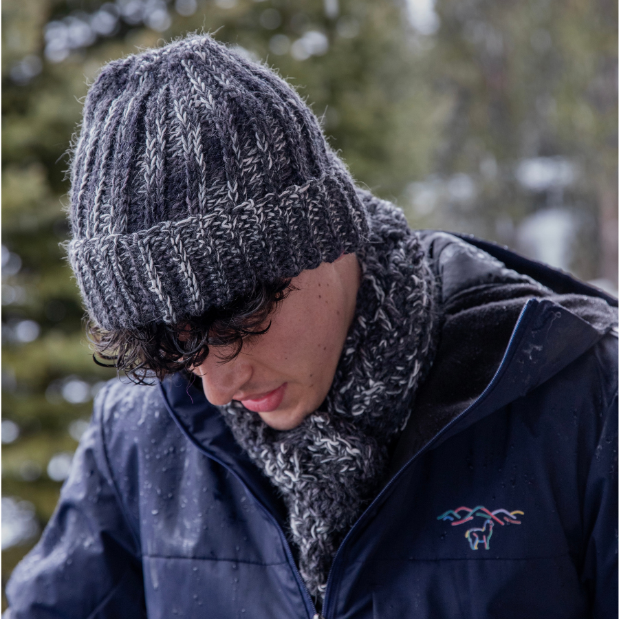 Close up of a young man with dark brown hair wearing Alpacas of Montana multi-gray knitted crochet handmade matching set of ridge ribbed hat and scarf with long tassels. Handmade items are a mixture of the colors black, charcoal, dark gray, light gray, and white.