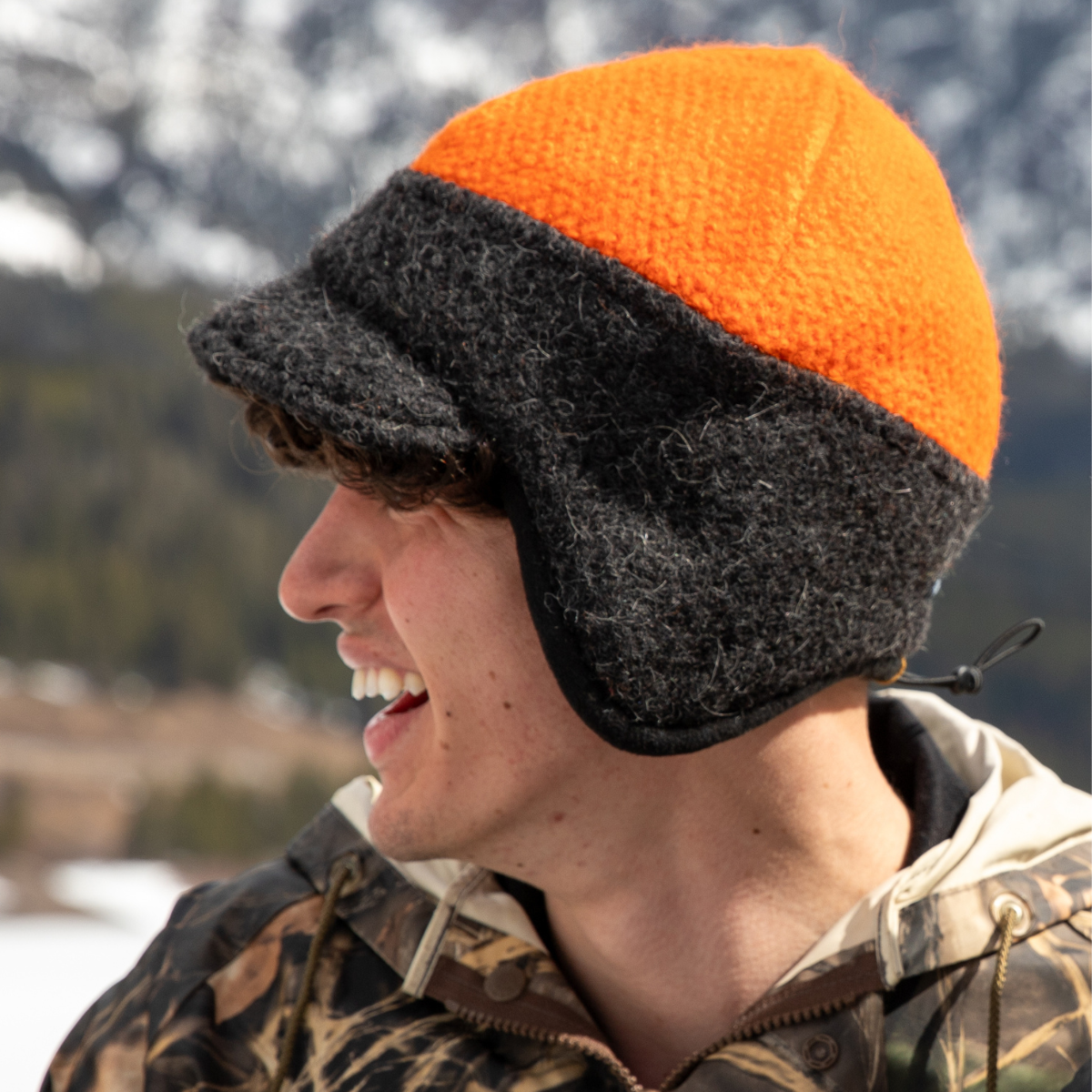 A close up photo of a young man wearing a hunting camouflage jacket and an orange and gray Alpacas of Montana an extremely warm cozy soft windproof comfortable moisture wicking thermal alpaca fleece wool windstopper winter hat for hiking, skiing, hunting, fishing, outdoors.