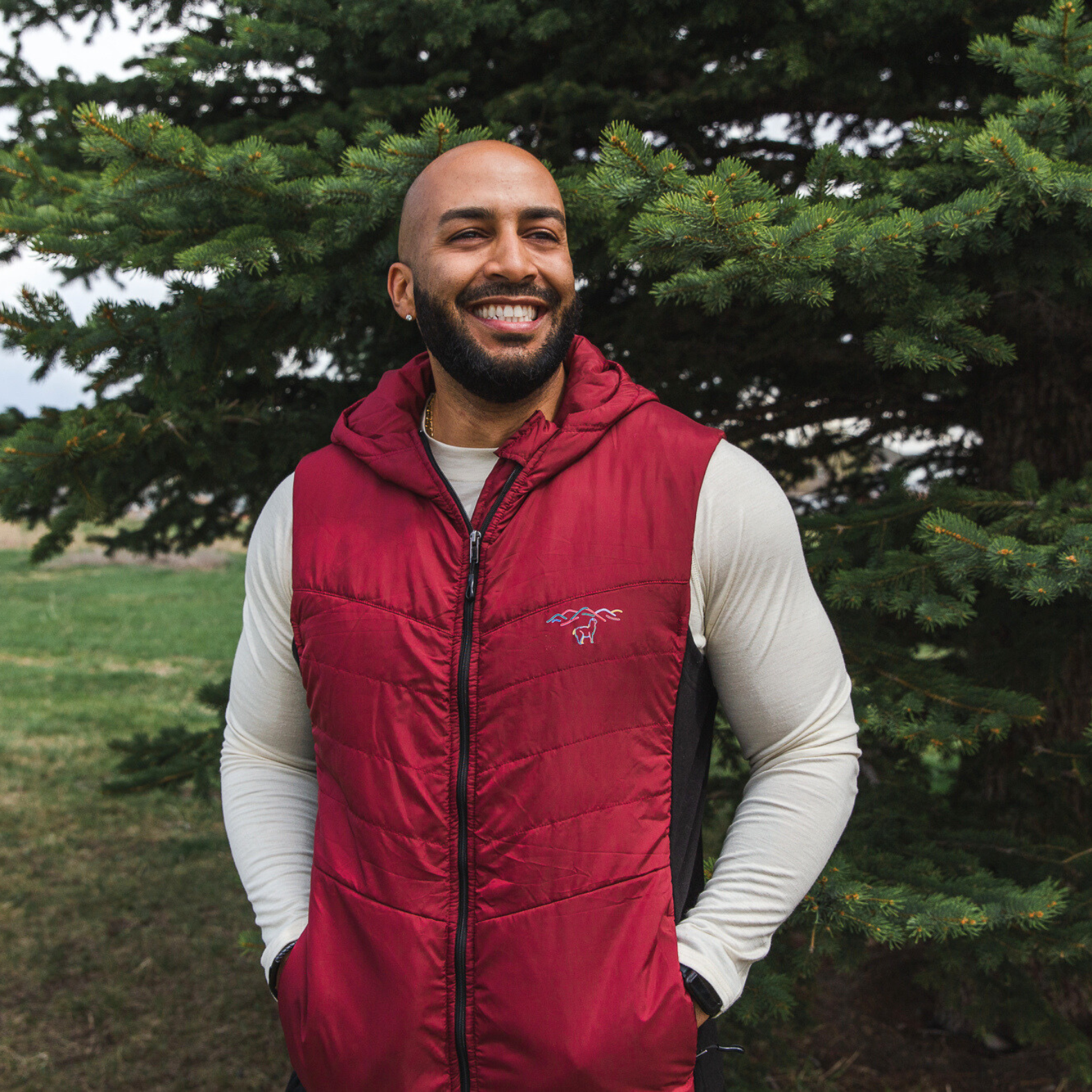 A smiling bald man with a black beard standing in front of a pine tree wearing an Alpacas of Montana natural white men's long sleeve performance tee and a cranberry red Alpacas of Montana lightweight athletic activewear outerwear breathable moisture wicking antimicrobial soft comfortable exercise alpaca men's stretch lite vest for running sports exercise work out hiking climbing camping biking