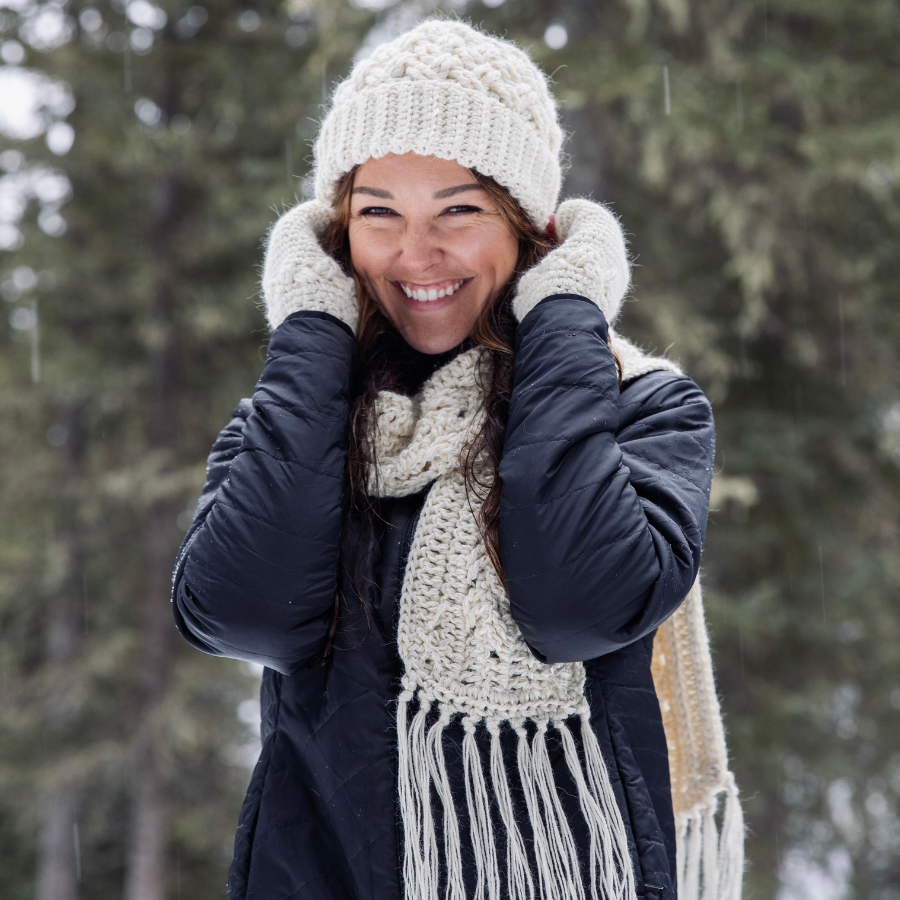 Close up of a woman with brown hair wearing Alpacas of Montana natural white knitted crochet handmade matching set of celtic pattern hat, scarf with long tassels on the end, and fingerless wristie mittens. Handmade items are all natural white color.