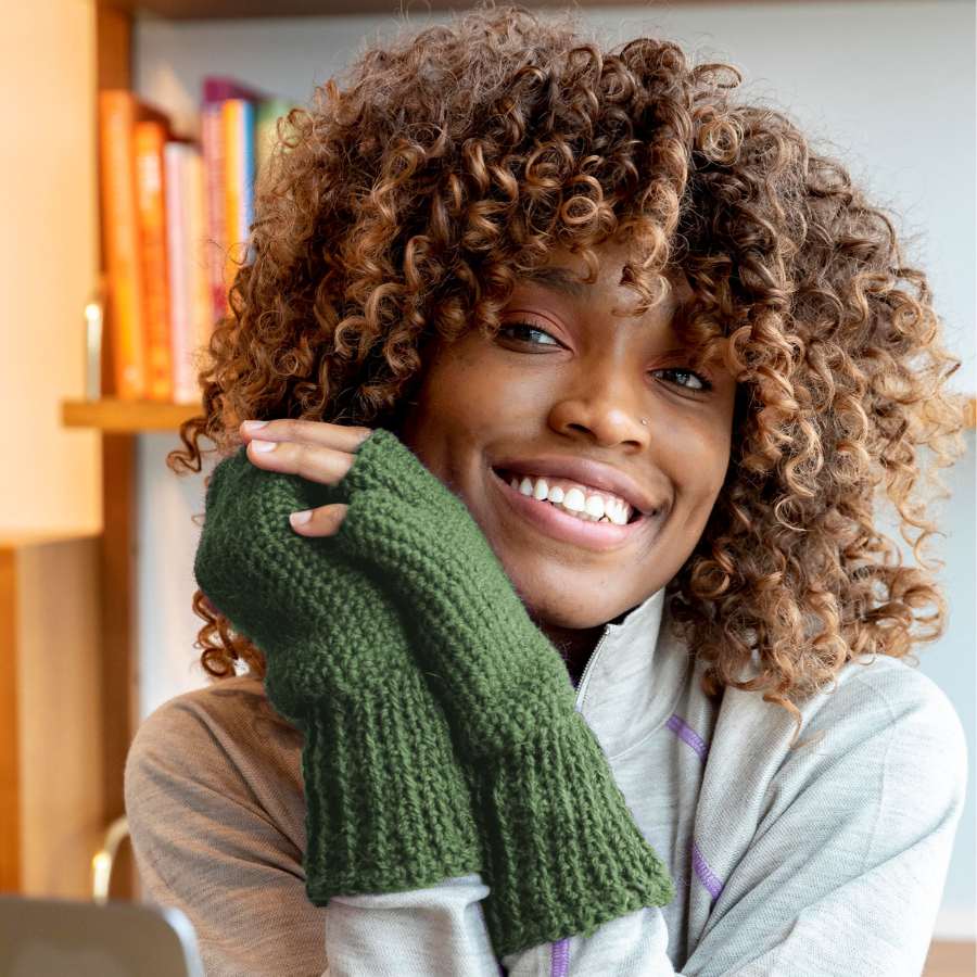 A close up photo of a smiling woman with curly brown hair wearing a pair of soft cozy comfortable fashionable moisture wicking knitted crochet fingerless gloves handmade in Montana from moss green alpaca wool yarn.