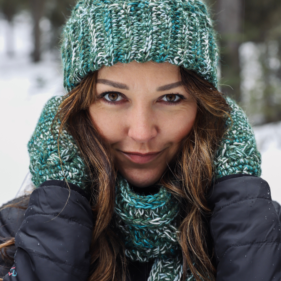 Close up of a woman with brown hair wearing Alpacas of Montana green knitted crochet handmade matching set of hat, scarf, and fingerless wristie mittens. Handmade items are a mixture of the colors moss green, teal, turquoise, seafoam, and white.