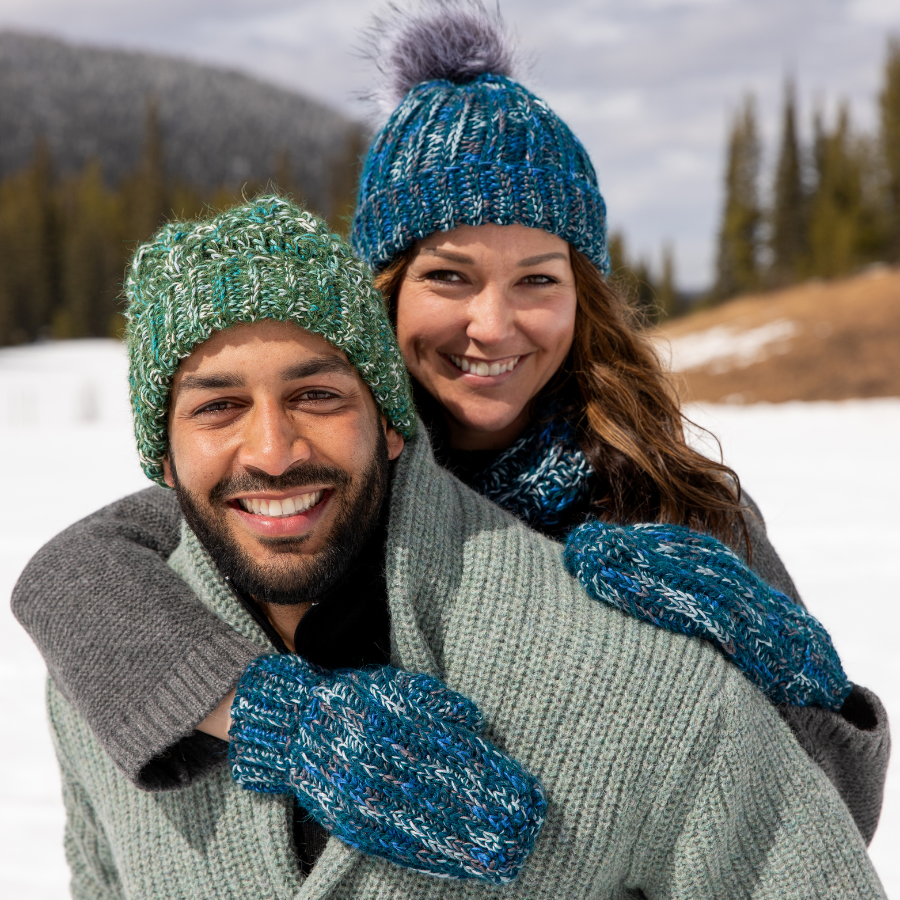 A smiling man with a black beard and a smiling woman with brown hair pose together in a winter scene. The man is wearing the Alpacas of Montana handmade knitted crocheted celtic hat in the colors moss green, teal, turquoise, seafoam, and white. The woman is wearing the Alpacas of Montana matching handmade set of ridge ribbed hat, scarf with tassels, and ridge ribbed mittens in the color ocean blue, dark turquoise, cobalt, cerulean, gray, and white.