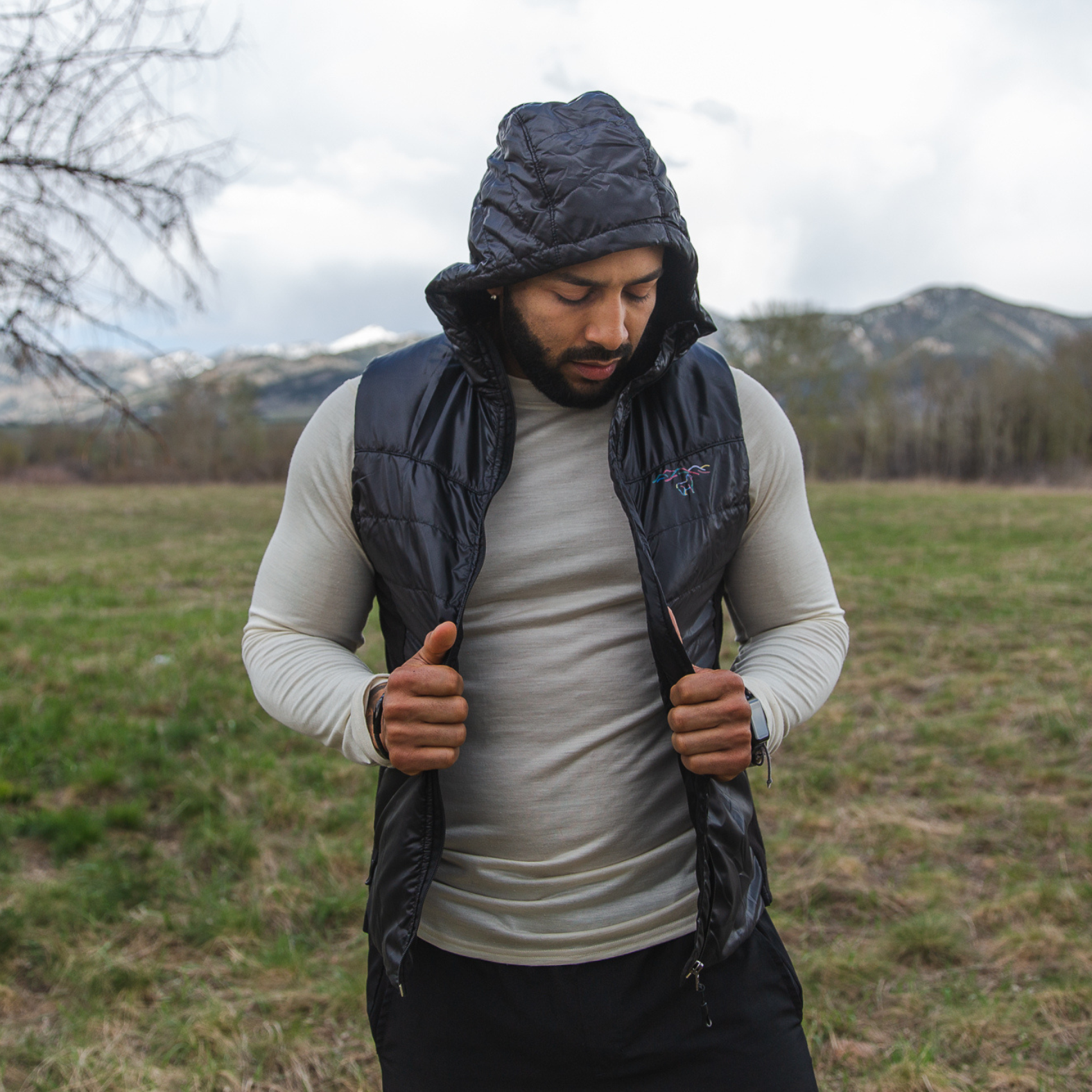 A man standing in a grassy field looking to the ground. He is wearing black pants, a natural white Alpacas of Montana men's long sleeve performance tee, and a black Alpacas of Montana lightweight athletic activewear outerwear breathable moisture wicking antimicrobial soft comfortable exercise alpaca men's stretch lite vest for running sports exercise work out hiking climbing camping biking