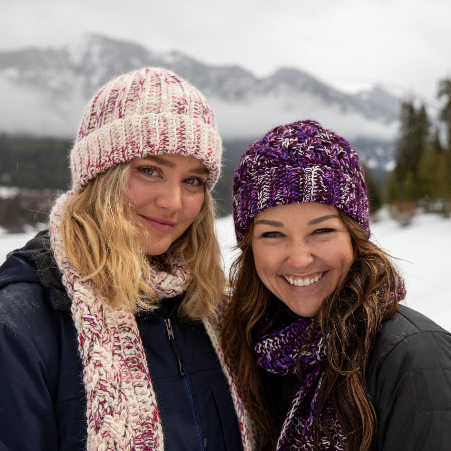 A smiling woman with blonde hair and a smiling woman with long brown hair pose in a winter scene side by side. The blonde woman is wearing the Alpacas of Montana handmade knitted crocheted ridge ribbed beanie and scarf with tassels in the colors fuchsia, dark pink, light pink, carnation, and white. The brown haired woman is wearing the Alpacas of Montana handmade celtic hat beanie and scarf with tassels in the colors deep purple, bright purple violet, and white.