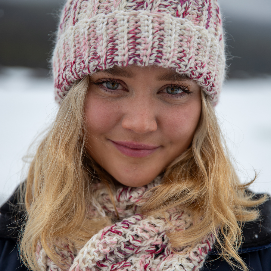 Close up photo of a smiling young blonde woman wearing the Alpacas of Montana handmade knitted crochet ridge ribbed hat and scarf with tassels made from yarn in the colors fuchsia, dark pink, light pink, carnation, and white.