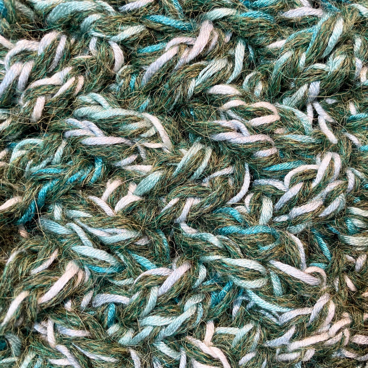 A close up photograph of the warm cozy handmade knit crochet Alpacas of Montana scarf, a mixture of the colors moss green, teal, turquoise, seafoam, and white. 