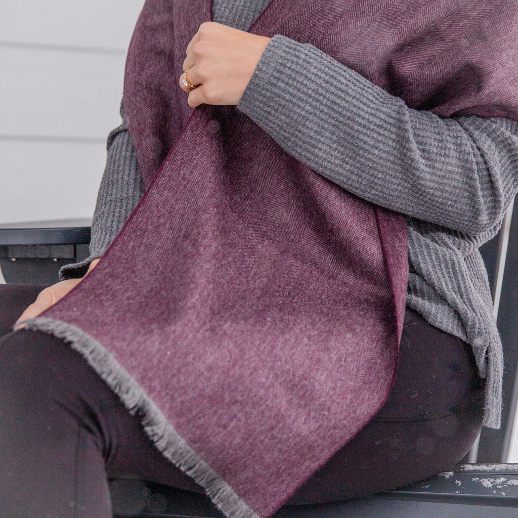 A close up photo of a person wearing black pants, a gray long sleeved shirt, and a soft cozy stylish comfortable women's fashion moisture wicking warm cute lovely herringbone patterned plum purple violet and gray alpaca wool scarf.