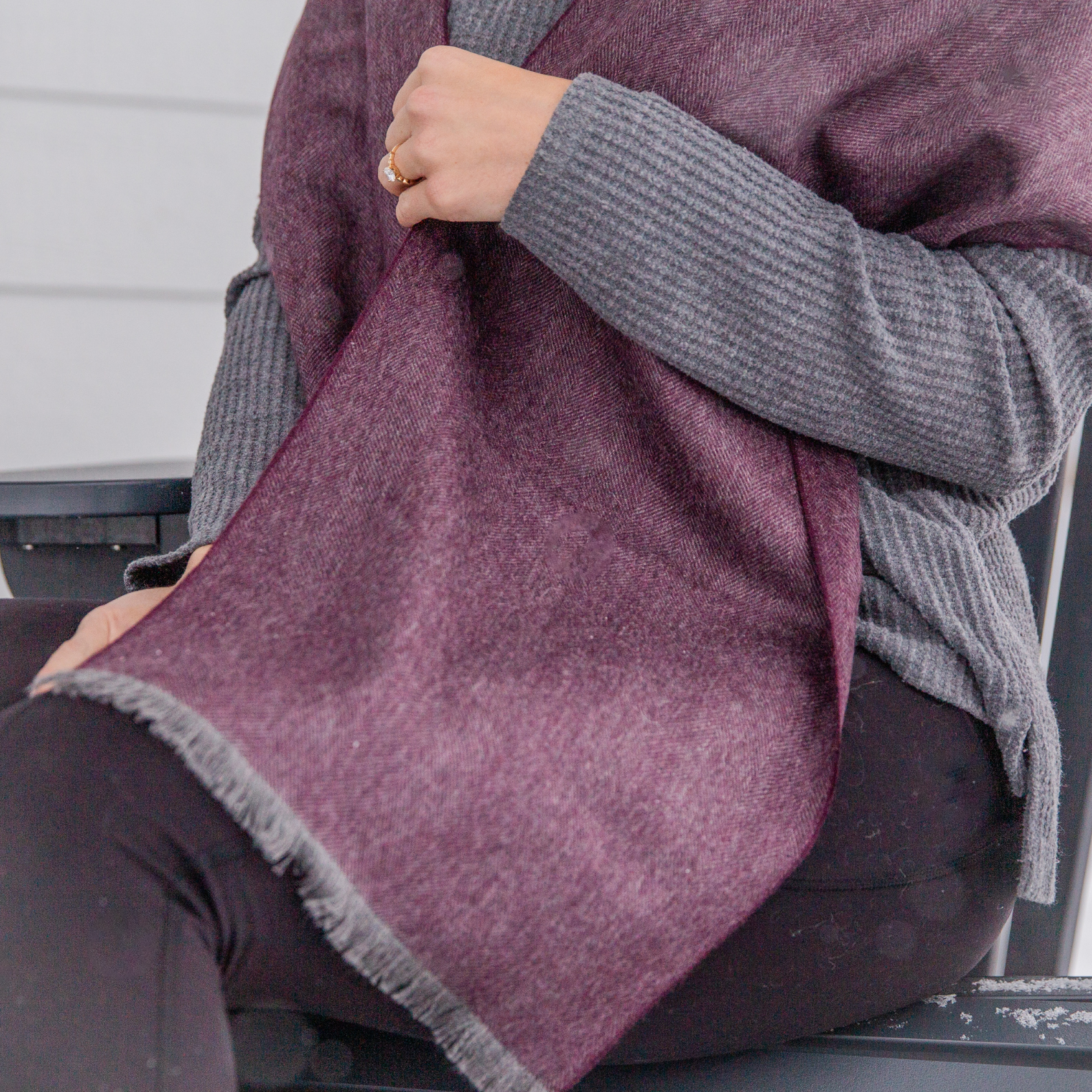 A close up photo of a person wearing black pants, a gray long sleeved shirt, and a soft cozy stylish comfortable women's fashion moisture wicking warm cute lovely herringbone patterned plum purple violet and gray alpaca wool scarf.