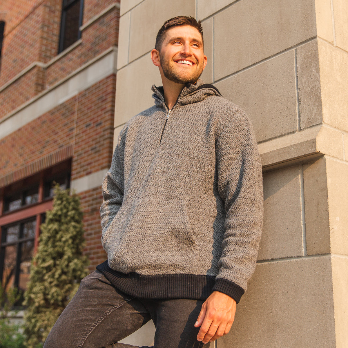 A smiling man with brown hair leaning against a beige brick wall on a sunny day. He is wearing black jeans and a light gray Alpacas of Montana thick warm cozy comfortable soft lined heavyweight warmth moisture wicking antimicrobial stylish men&#39;s fashion oversized alpaca wool hoodie.