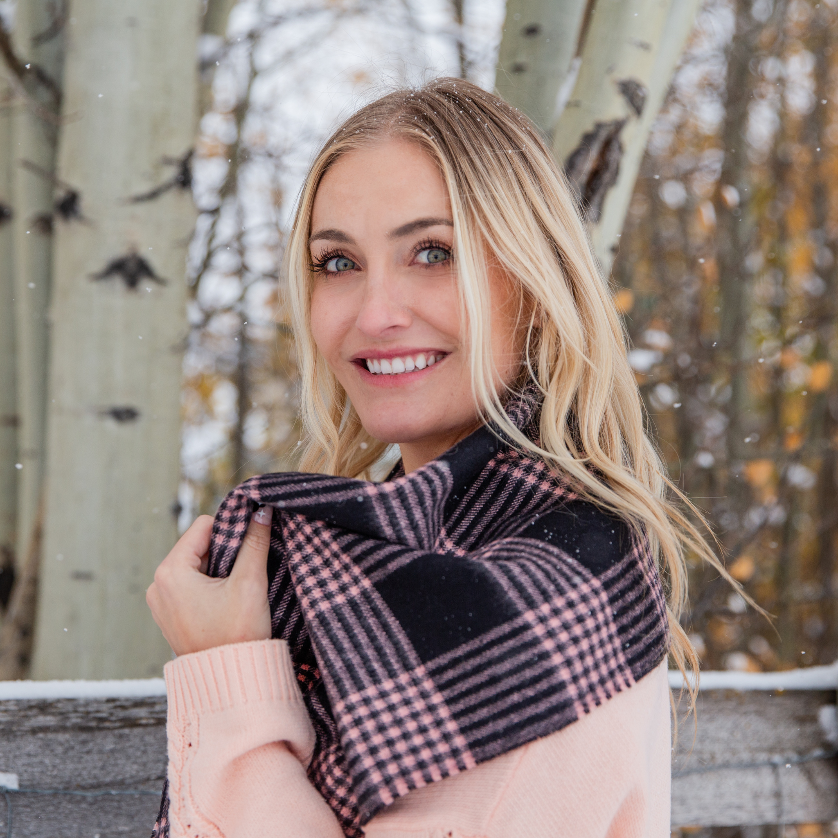 A smiling blonde haired woman standing against a fence with aspen trees and snow in the background. She is wearing a pink sweater and an Alpacas of Montana soft stylish women's fashion comfortable cozy cute warm alpaca wool blush pink and black plaid pattern scarf with tassels.