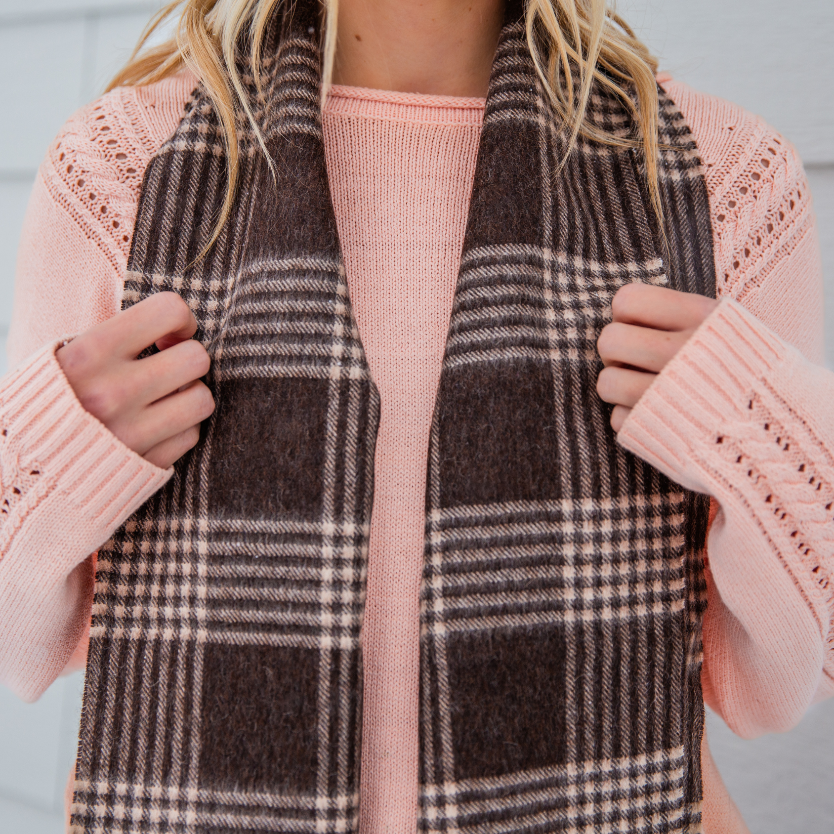A close up photo of a woman in a pink woven sweater and an Alpacas of Montana soft stylish women's fashion comfortable cozy cute warm alpaca wool latte brown and chocolate brown plaid pattern scarf with tassels.