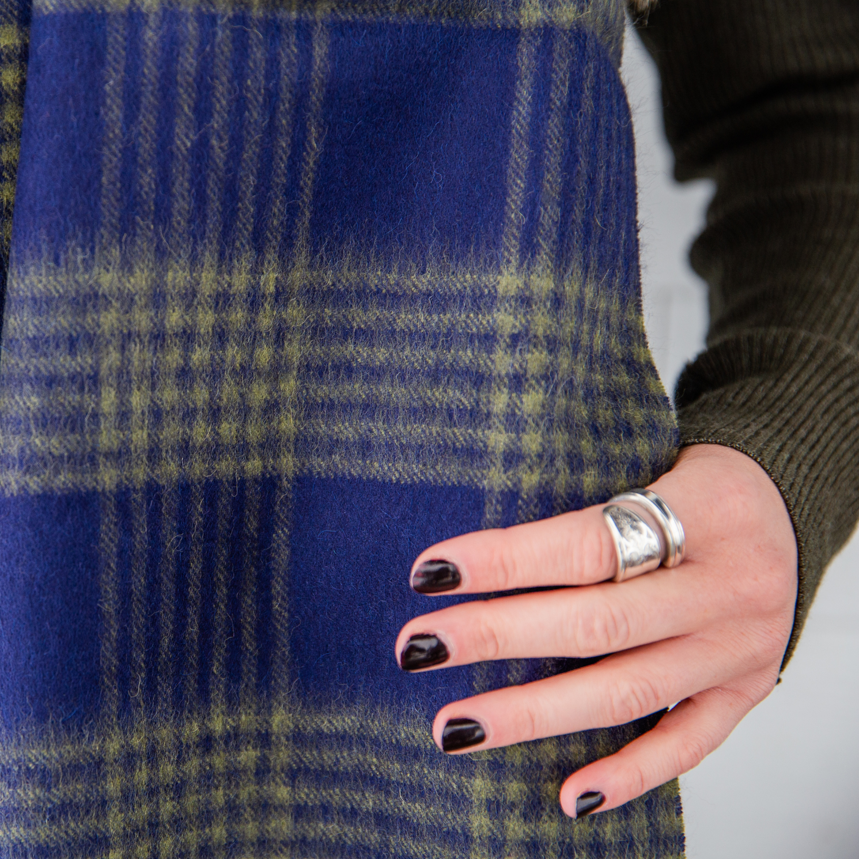 A close up photo of a woman's hand with black painted nails holding an Alpacas of Montana soft stylish women's fashion comfortable cozy cute warm alpaca wool green and navy blue plaid pattern scarf with tassels.