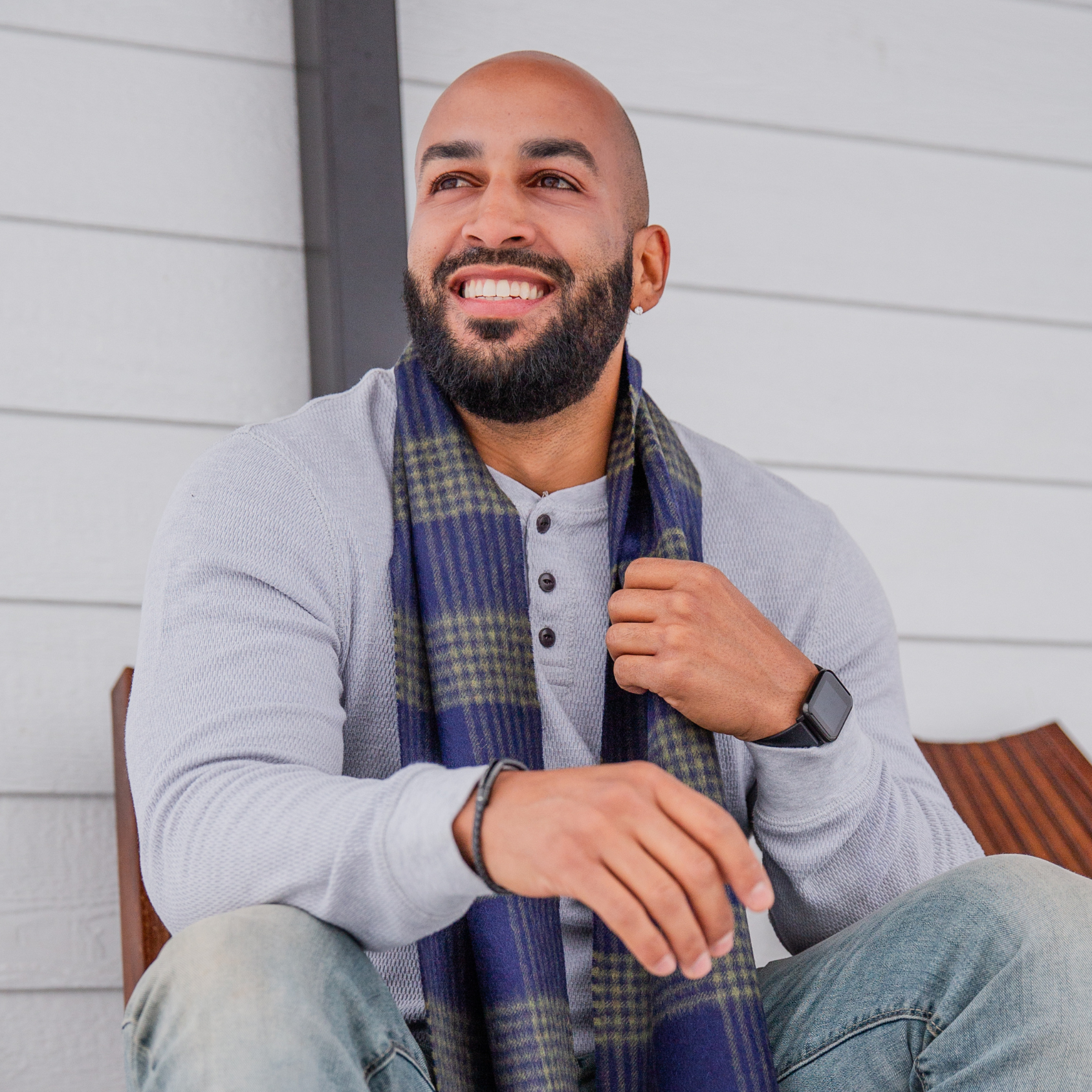 A smiling bald man with a black beard sitting on a wooden chair and looking into the distance with a white wall behind him. He is wearing blue jeans, a gray long sleeve shirt, and an Alpacas of Montana soft stylish men's fashion comfortable cozy cute warm alpaca wool green and navy plaid pattern scarf with tassels.