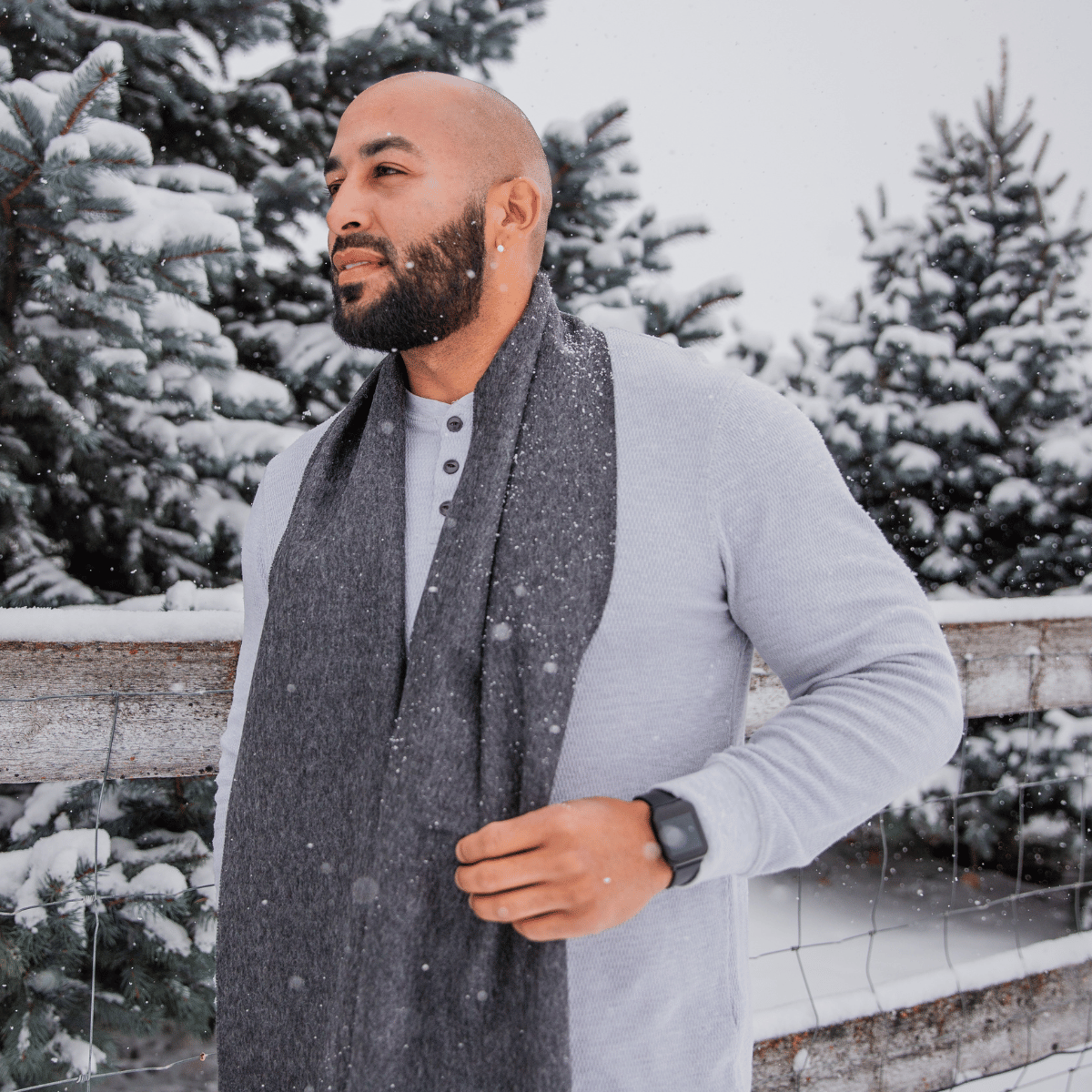 A bald man with a black beard looking into the distance standing in front of a wooden fence and pine trees. He is wearing a light gray long sleeve shirt and an Alpacas of Montana soft stylish women's fashion comfortable cozy cute warm alpaca wool dark gray color solid woven scarf with tassels.