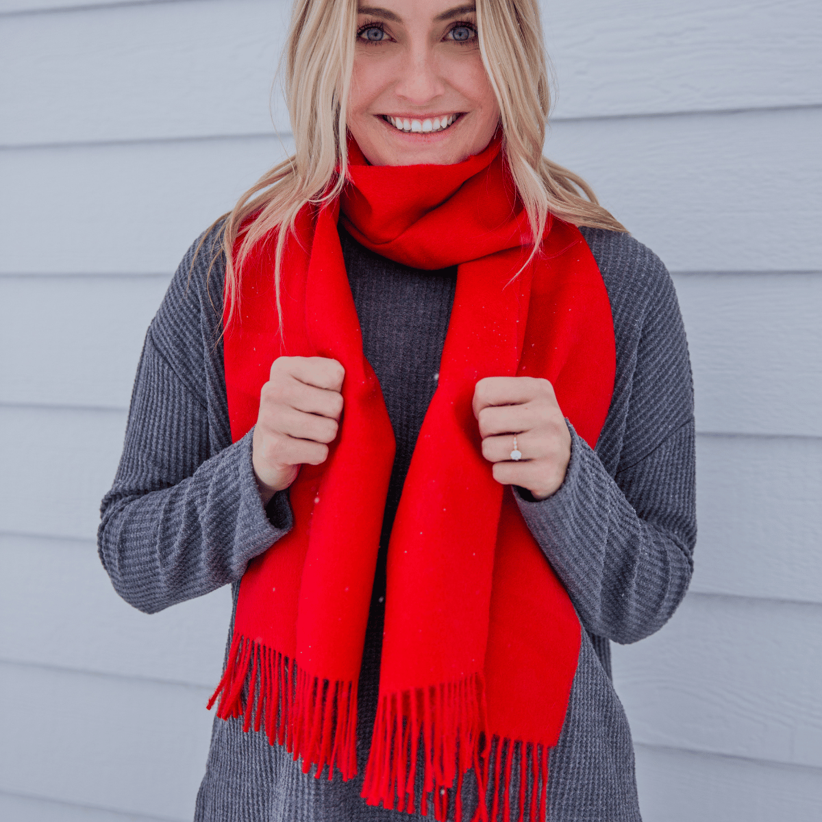 A blonde woman smiling at the camera and wearing a long sleeve dark gray shirt and an Alpacas of Montana soft stylish women's fashion comfortable cozy cute warm alpaca wool vibrant scarlet red solid woven scarf with tassels.