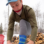 A man leaning over to tie his shoes in front of a wood pile. He is wearing Alpacas of Montana blue mid-crew hiking sock, gray long johns, an olive green Alpacas of Montana men's base layer top, and a lightweight cozy comfortable moisture wicking warm breathable all seasons thin thermal outerwear skiing hiking climbing outdoors hunting fishing high desert neck gaiter made from scarlet red and light camel brown alpaca.