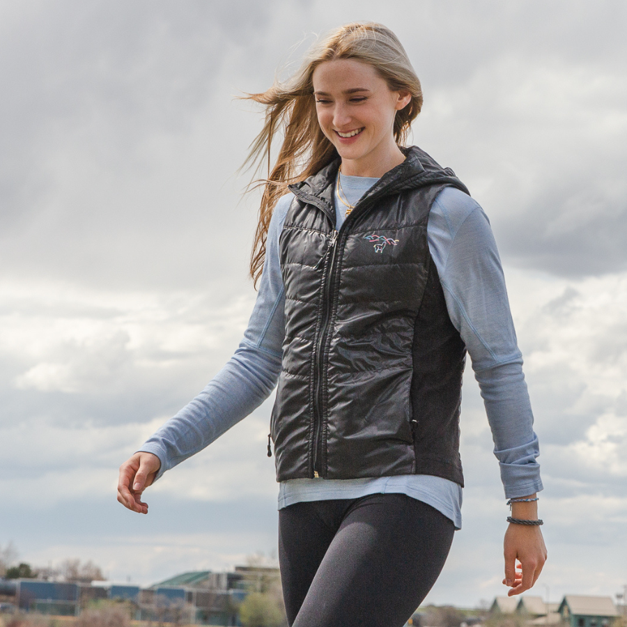 A blonde woman walking in gray leggings, a light blue long sleeve shirt, and a black Alpacas of Montana lightweight athletic activewear outerwear breathable moisture wicking antimicrobial soft comfortable exercise alpaca women's stretch lite vest for running sports exercise work out hiking climbing camping biking