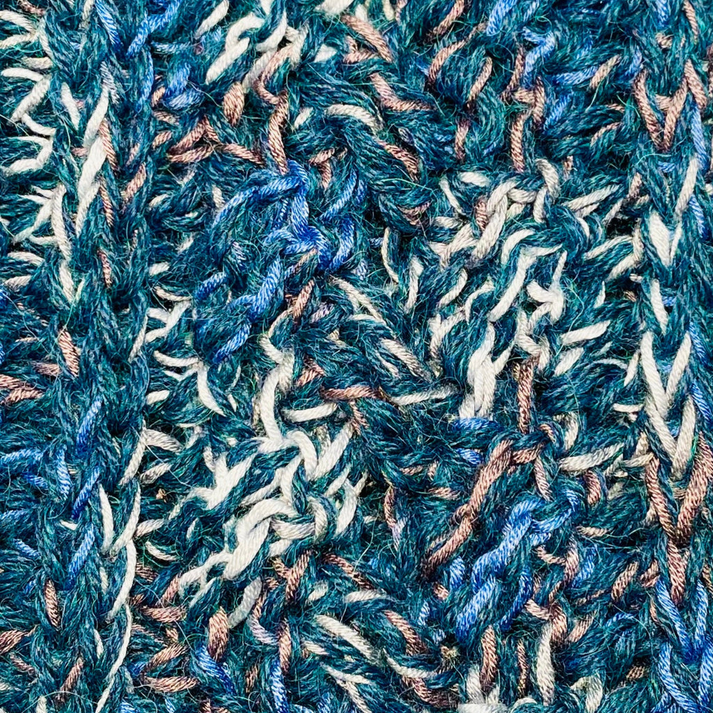A close up photograph of the warm cozy handmade knit crochet Alpacas of Montana scarf, a mixture of the colors ocean blue, dark turquoise, cobalt, cerulean, gray, and white. 